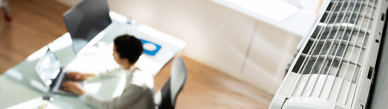 AndreyPopov/iStock Blick von der oberen Zimmerdecke am Klimagerät vorbei auf eine Person am Schreibtisch sitzend.